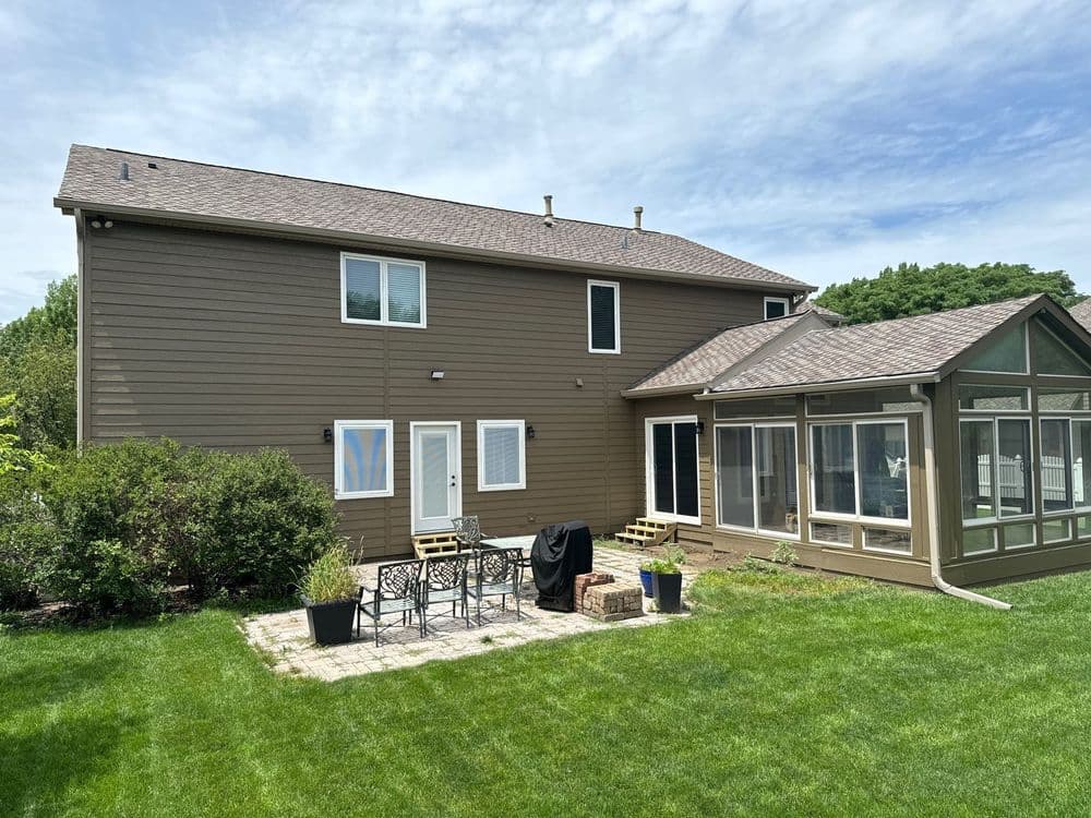 Brown two-story house with a patio and sunroom, surrounded by green grass and shrubs.